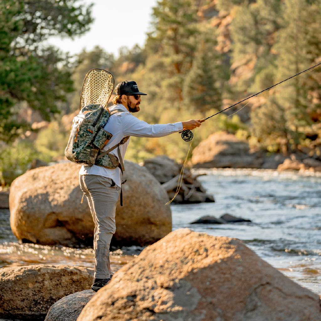 man fly fishing next to river in green long sleeve pearl snap