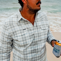 Man holding a drink on the beach wearing the blue and grey flannel