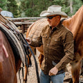 man putting saddle on horse in the mountains wearing brown corduroy